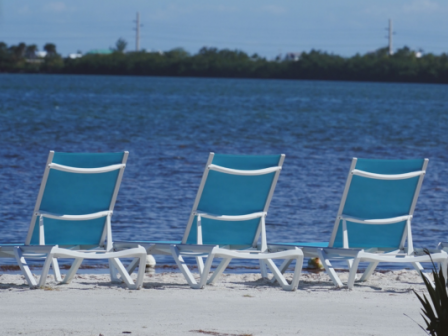 Three blue beach chairs on a sandy shore overlook a calm body of water, with a line of trees in the distance under a clear blue sky.