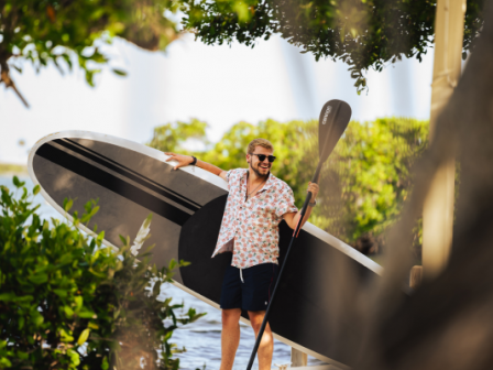 A person stands by water, holding a paddleboard and paddle, surrounded by greenery, wearing sunglasses and a patterned shirt.