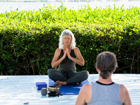 Two people practicing yoga outdoors, one meditating on a mat with a sound bowl nearby, surrounded by greenery and a peaceful background.