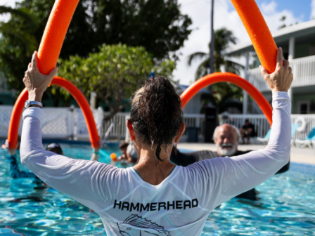 People are in a pool, using orange pool noodles for exercise or fun. A person wears a shirt with "HAMMERHEAD" printed on the back.