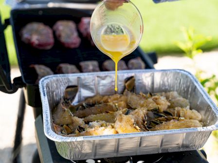 Grilling scene with butter being poured over seafood in a tray; steaks are on the grill in the background.