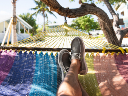 Person relaxing in a colorful hammock under a tree, wearing grey shoes, with a tropical background of houses and palm trees.