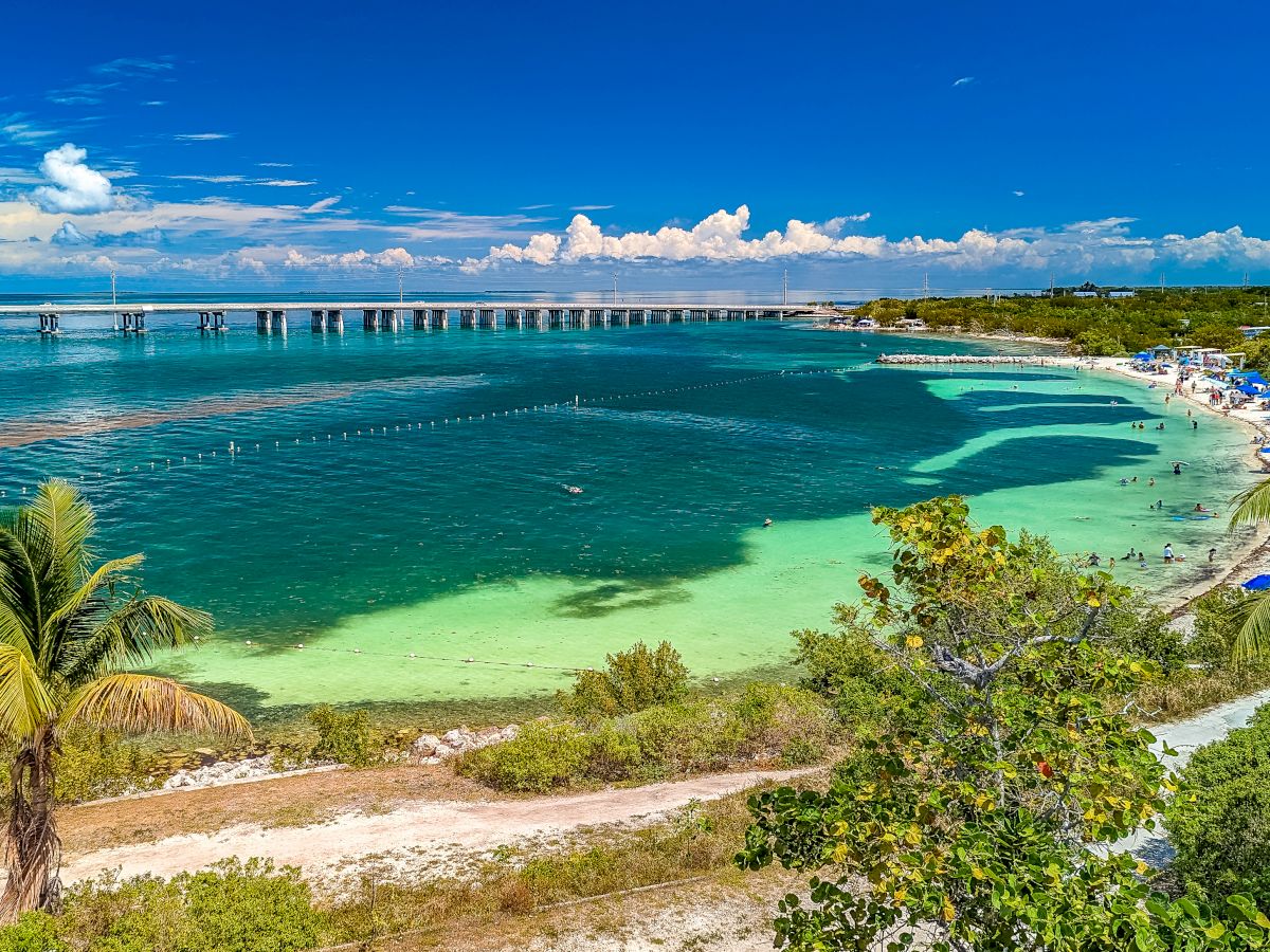 A tropical beach scene with clear turquoise water, palm trees, and a bridge in the background under a blue sky with scattered clouds.