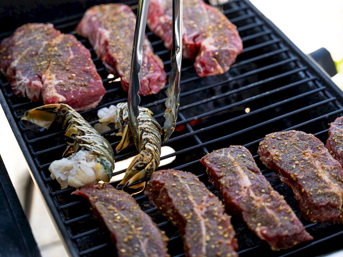 Grilling steaks and lobster tails on a barbecue with tongs, seasoned with spices.