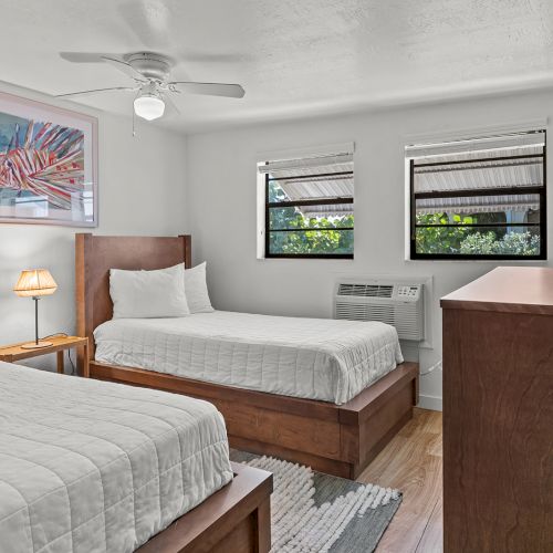 The image shows a bedroom with two beds, a dresser, a small lamp, and a fish painting on the wall, with windows letting in natural light.