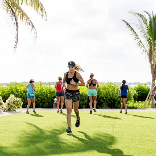 A group of people exercising outdoors on a grassy area surrounded by tropical plants and palm trees, under a partly cloudy sky.