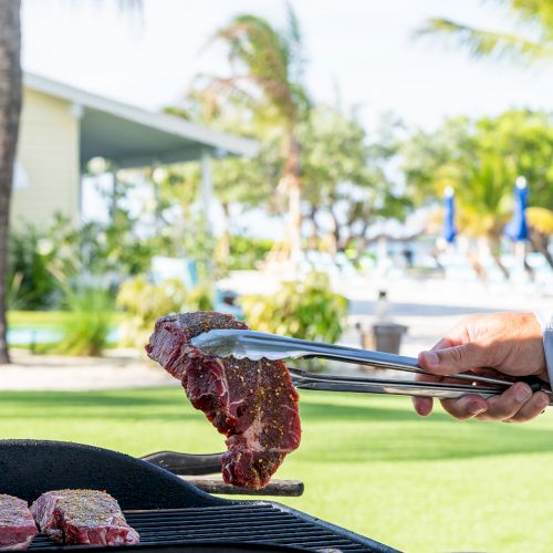 A person is grilling steaks outdoors with tongs on a sunny day near a house and trees in the background.