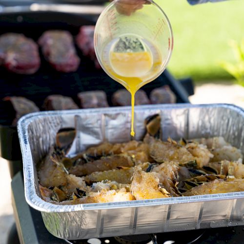 Melted butter is being poured over lobster tails in a foil tray, while meat cooks on the grill in the background.