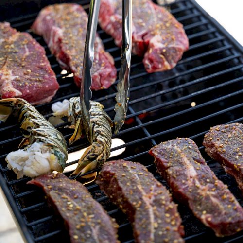 The image shows seasoned meat and lobster tails being grilled on an outdoor barbecue.