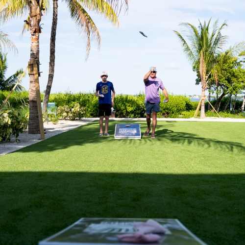 Two people are playing cornhole on a grassy area near palm trees and a pool, with a scenic backdrop of trees and water.