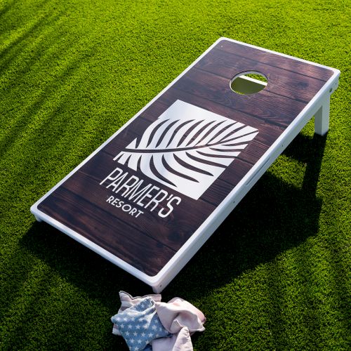 A cornhole board with a leaf design and text, on a grass surface, with bean bags nearby.