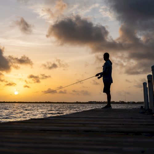 A person is fishing on a dock at sunset, silhouetted against a colorful sky and calm water.