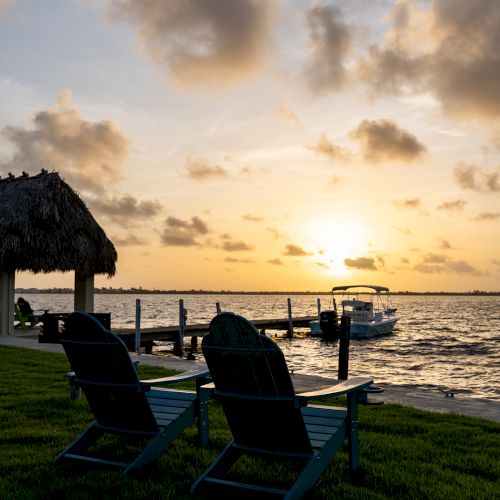 Two chairs face a tranquil waterfront at sunset, with a thatched-roof structure and boat adding to the serene scene.