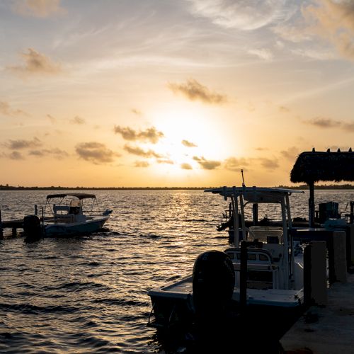 A serene sunset over a waterfront with boats docked, a thatched-roof shelter, and a calm, rippling water surface completes the scene.