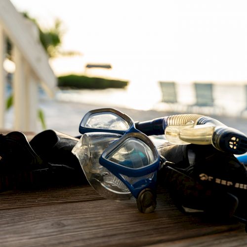 A pair of diving goggles and a snorkel rest on a wooden deck with a blurred beach scene in the background.