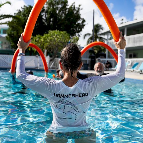 A person in a pool holds two orange pool noodles, wearing a shirt with text on the back, with others present in a sunny outdoor setting.