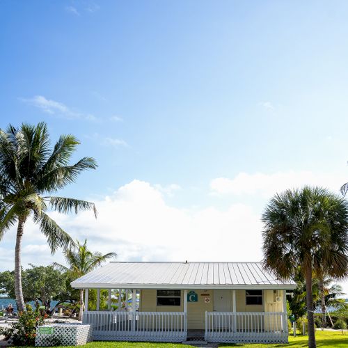 A small white house with a metal roof, surrounded by lush green lawn and palm trees, under a clear blue sky with scattered clouds.