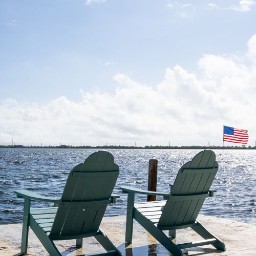 Two chairs face a body of water under a blue sky, with an American flag in the background, creating a peaceful scene.