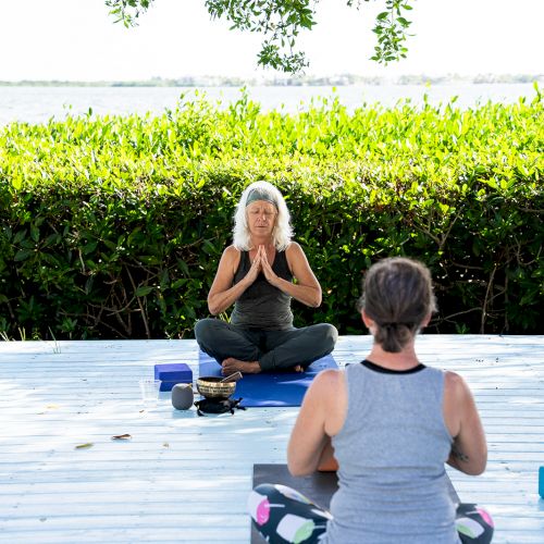 Two people are practicing yoga outdoors on a wooden deck with greenery and a water view in the background.