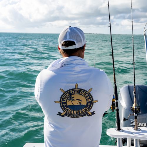 A person in a white shirt with a fishing logo stands on a boat with fishing rods, overlooking the sea under a partly cloudy sky.