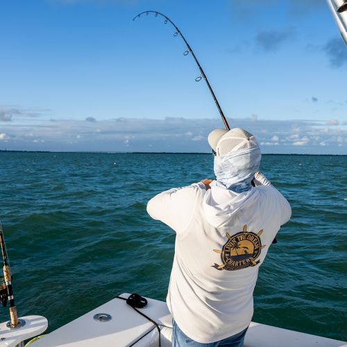 A person is fishing from a boat in the ocean, wearing a hat and shirt, with a fishing rod bent by a catch.