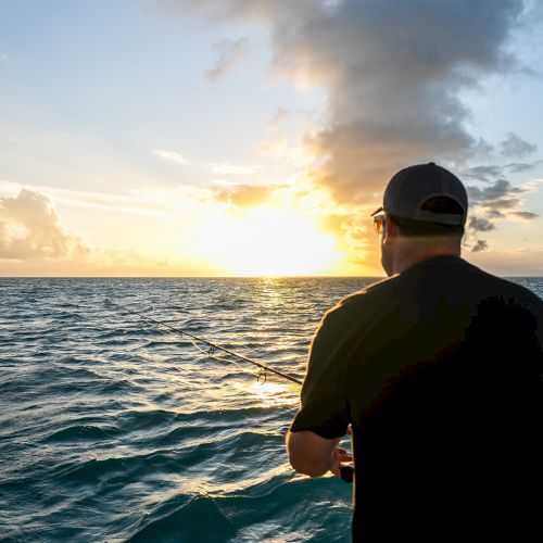 A person in a cap stands on a boat, looking at a serene ocean sunset with scattered clouds in the sky.