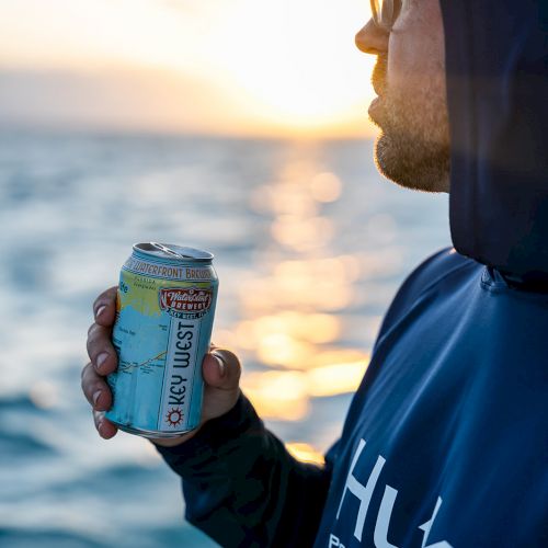 A person in a hoodie holding a can, standing near water at sunset.