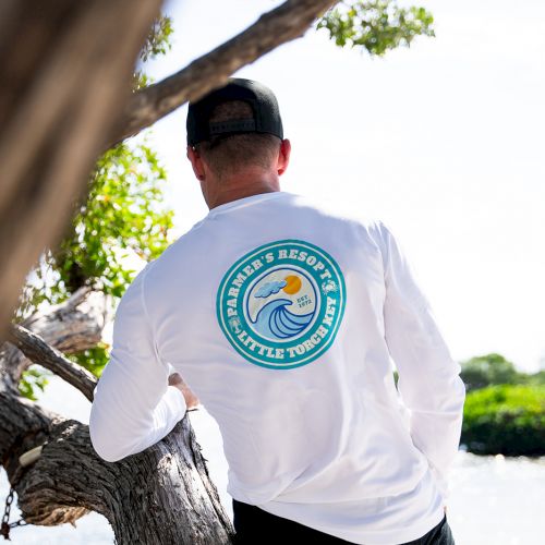 A person wearing a white long-sleeve shirt with a circular logo on the back, leaning on a tree by the water.