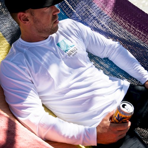 A person relaxes in a colorful hammock, wearing a white shirt and hat, holding a can of drink, with sunlight casting shadows.