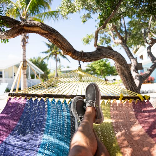 A person relaxing in a colorful hammock under trees, with legs extended and sneakers visible, enjoying a sunny day outdoors.