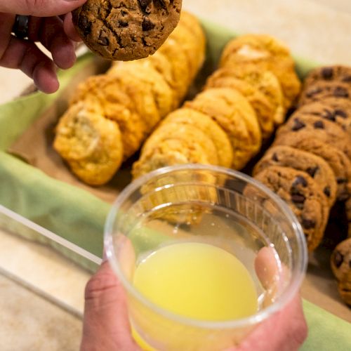 A person holds a chocolate chip cookie and a cup of yellow juice, with trays of assorted cookies in the background.