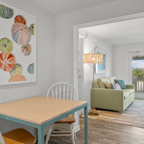 The image shows a cozy room with a small dining table and chairs, a living area with a green sofa, and decorative wall art, leading to a balcony.