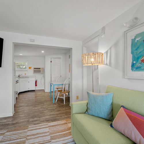 A cozy living room with a green sofa, colorful cushions, wall art, TV, and view into a kitchen with white cabinetry and a small dining area.