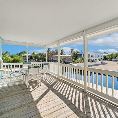 A spacious porch overlooks a serene waterway, featuring outdoor seating and bright skies, surrounded by palm trees and neighboring houses.