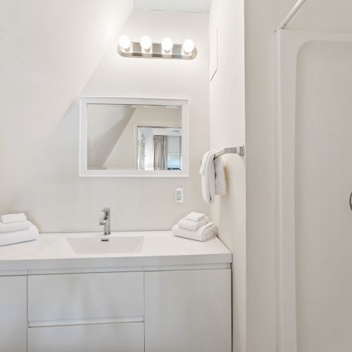 A modern bathroom with a white vanity, mirror, stacked towels, and a shower; bright lighting enhances the clean design.