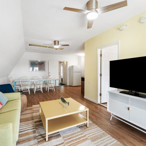 A cozy living room with a green sofa, colorful cushions, coffee table, large TV, dining area, ceiling fans, and wooden flooring.