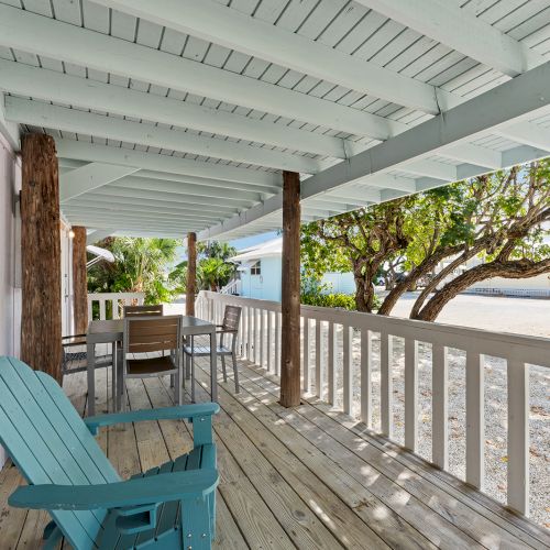 A wooden porch with light blue decor, a table with chairs, and a relaxing chair overlooks a sandy area with trees.