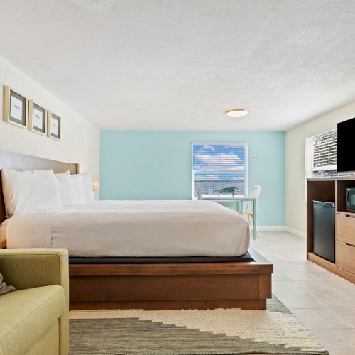 A modern hotel room with a bed, sofa, large TV, mini fridge, and a window showing a view outside with blue skies and clouds.