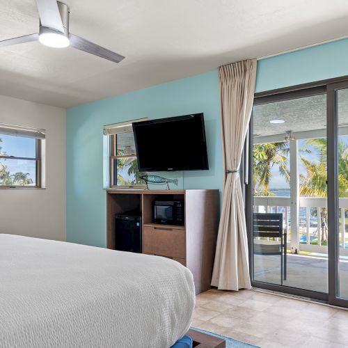 The image shows a hotel room with a bed, wall-mounted TV, microwave, and a view of a balcony with palm trees and ocean scenery.