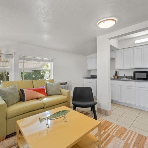 A cozy living room with a yellow couch, modern lamp, coffee table, and small kitchen area with appliances.