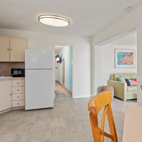 The image shows a small kitchen with white cabinets, a fridge, stove, and some furniture. A hallway and living area are in the background.