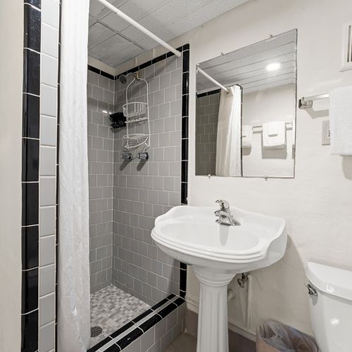 A small bathroom with a shower, pedestal sink, toilet, mirror, and towel rack. Towels are neatly stacked on a shelf.