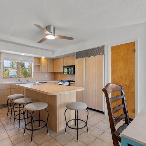 A modern kitchen with wooden cabinets, a central island with stools, a ceiling fan, and a glimpse of a living area.