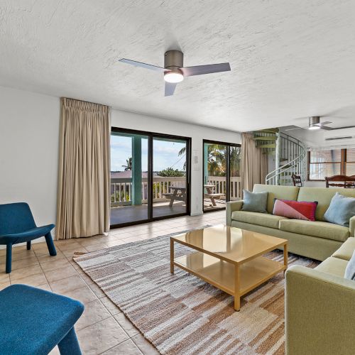 A modern living room with a TV, blue chairs, green sofas, a wooden coffee table, and a view of an outdoor area through glass doors.