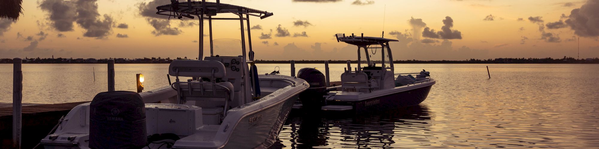 Two boats are docked on calm waters under a golden sunset sky, with silhouetted clouds and a serene horizon in the background.