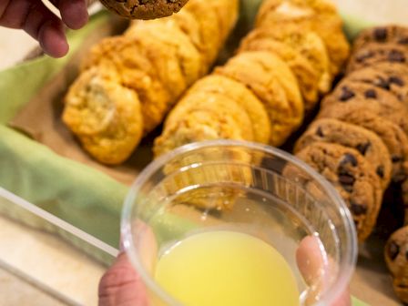 A person holds a chocolate chip cookie and a cup of yellow juice, with a tray of assorted cookies in the background.