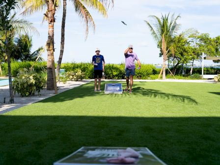 Two people play cornhole on a grassy area with palm trees and a pool nearby.