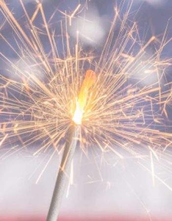 A lit sparkler with bright sparks is in the foreground, and a blurred background resembles the U.S. flag.