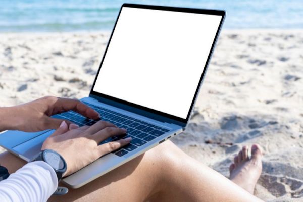 A person is sitting on a sandy beach using a laptop with a blank screen, overlooking the ocean under a clear sky.