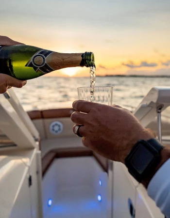 A person pours champagne into a glass on a boat at sunset, with the ocean in the background.